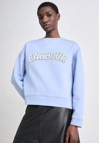 Világoskék kapucnis pulóver fehér "BARBOUR" hímzéssel, laza szabású, kerek nyakkivágású, bordás mandzsettákkal; fekete bőr szoknyával viselve.