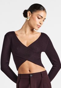 Top lungo a maniche con costine color borgogna, scollo a V e design incrociato sul davanti, abbinato a pantaloni a vita alta. Il tessuto ha una texture liscia.