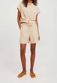 Beige kortärmad skjorta med krage och ficka fram, ihop med beige shorts med elastisk midja. Bärs med gula flip-flops.