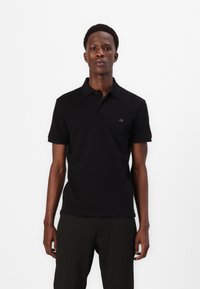 Homem a usar uma camisa polo preta de manga curta com um pequeno logótipo no peito e calças escuras com riscas verticais, de pé contra um fundo branco.