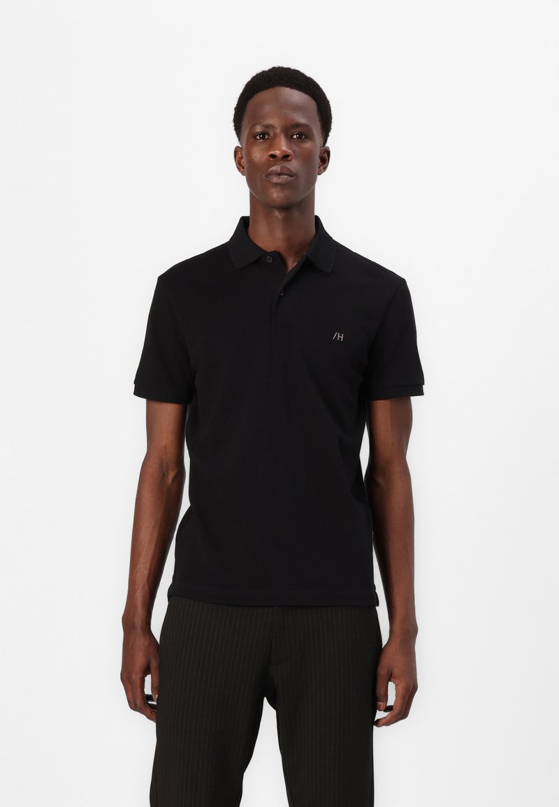 Homem a usar uma camisa polo preta de manga curta com um pequeno logótipo no peito e calças escuras com riscas verticais, de pé contra um fundo branco.