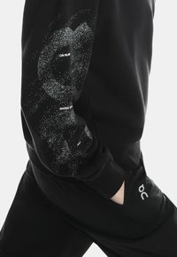 Fekete pulóver, amelyen a bal ujján fehér pöttyös textúrájú minta található. Tartalmazza az "ON RUN" és a "SWISS ENGINEERING" logót.