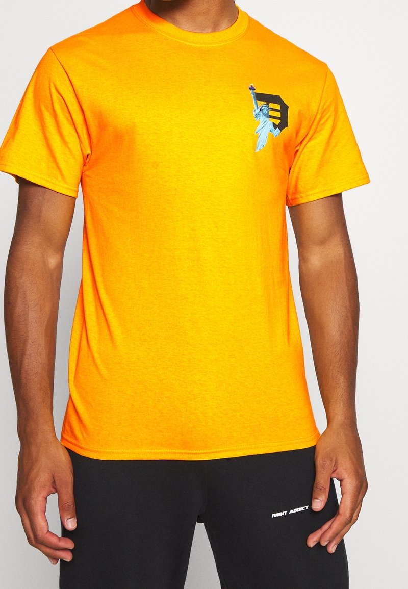 Homme portant une chemise orange vif à manches courtes avec un petit motif de la Statue de la Liberté sur la poitrine, associée à un pantalon noir.