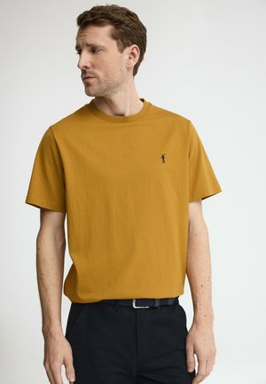 Hombre con camiseta de manga corta amarillo mostaza con pequeño logo negro de golfista, y pantalones negros con cinturón tejido, mirando hacia su izquierda.