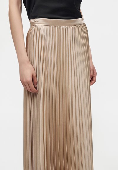 Femme portant une jupe plissée métallique dorée avec une ceinture en satin et un haut noir, les mains reposant le long du corps.