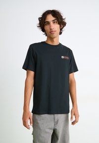 Sort bomulds t-shirt med korte ærmer, rund halsudskæring og lille rektangulært rødt og hvidt logo på venstre bryst. Modellen har lysegrå bukser på.