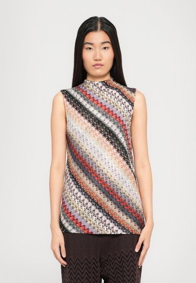 Jeune femme aux cheveux raides noirs portant un haut sans manches à col montant avec des rayures diagonales multicolores et un pantalon à motifs zigzag noirs.
