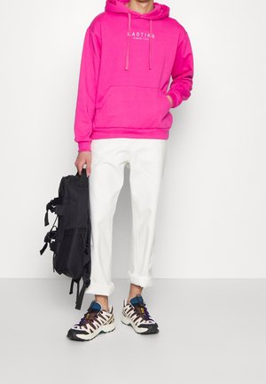 Sweat à capuche rose avec poche avant, pantalon blanc fuselé, et sac à dos noir. Les chaussures présentent un motif en crème, marron et accents bleus.