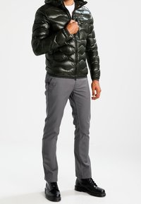 Grüne Stepp-Winterjacke mit Kapuze, gestepptem Muster und Frontreißverschluss. Kombiniert mit grauer Hose und schwarzen Schnürstiefeln.