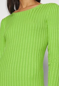 Top lungo a maniche lunghe verde a costine con scollo a barca. Il tessuto presenta un motivo testurizzato di strisce verticali, che mette in risalto la sua silhouette aderente.
