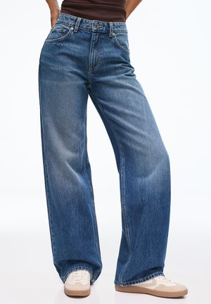 Jean droit en denim bleu porté avec des baskets blanches et un top marron, montré de la taille aux pieds sur un fond uni.