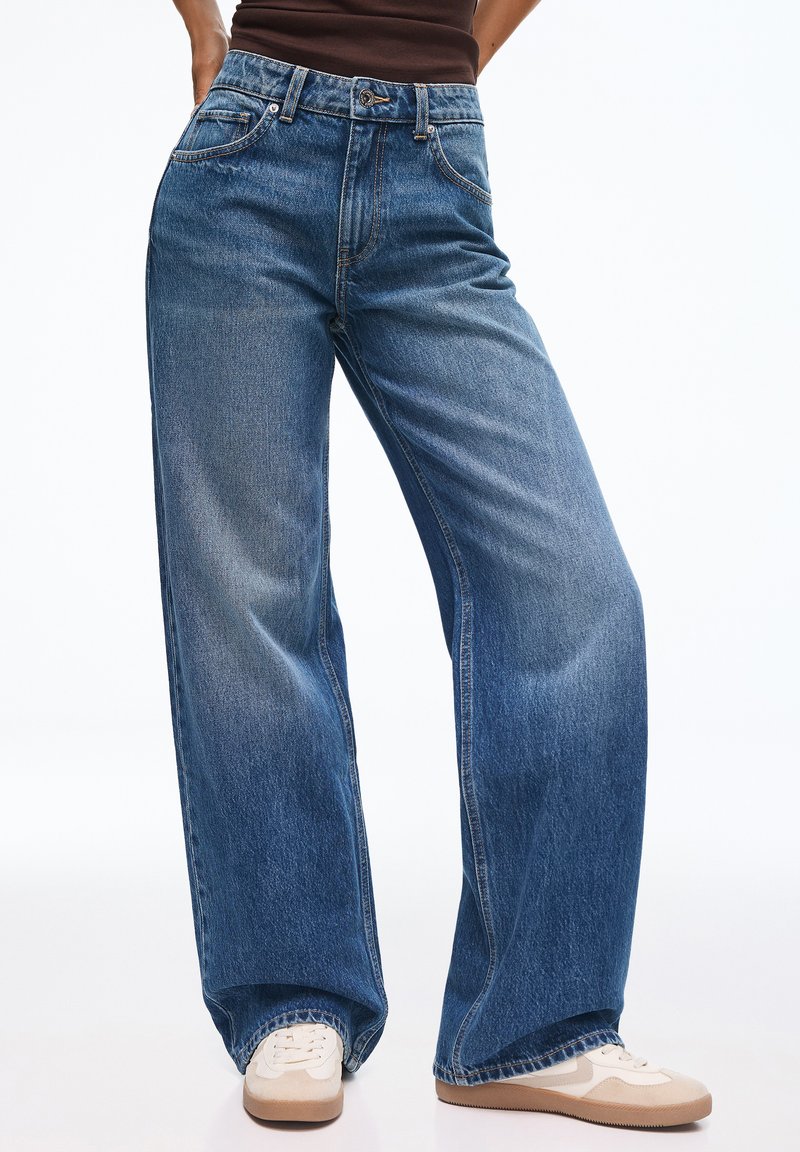 Jean droit en denim bleu porté avec des baskets blanches et un top marron, montré de la taille aux pieds sur un fond uni.
