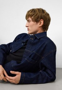 Jeansjacka i mörkblått med krage, framfickor och kontrasterande sömmar. Modellen bär en svart skjorta under. Platt yta i bakgrunden.