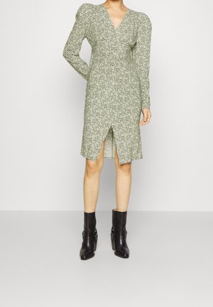 Femme portant une robe verte à fleurs longueur genou avec manches longues, fente frontale, et des bottines noires, debout devant un fond clair.