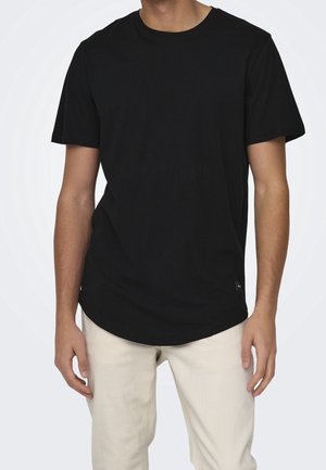 Homme portant un t-shirt noir uni à manches courtes et col rond, ainsi qu'un pantalon beige clair, sur un fond blanc.