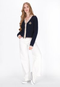Marineblaue Strickjacke mit Kabelmuster, weißen Akzenten und goldenen Knöpfen. Enthält ein Logo-Patch auf der Brust, kombiniert mit weißen weiten Hosen und Sneakers.