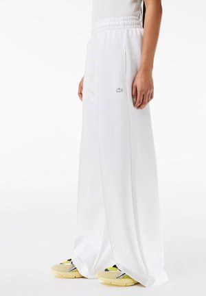 Personne portant un pantalon blanc ample avec une taille élastique et le logo Lacoste, assorti à des baskets jaunes et beiges sur fond blanc.