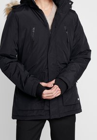 Svart isolerad jacka med huva, med fuskpälsdetaljer, dragkedjefickor på bröstet och elastiska ärmslut. Mjuk textur och slitstarkt material.