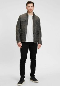 Dunkelgraue, texturierte, knöpfbare Jacke mit vier Fronttaschen, getragen über einem weißen Hemd, kombiniert mit schwarzen Jeans und schwarzen Sneakers.