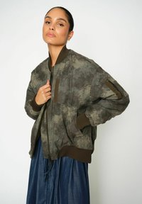 Maslinasto zelena bomber jakna s teksturiranim uzorkom, rebrastim manžetama i džepovima. Tkanina izgleda meko i lagano sjajno.