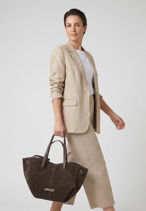 Zelfverzekerde vrouw in beige pak en wit overhemd draagt een grote donkerbruine suède draagtas met het label "Venezia" tegen een effen achtergrond.