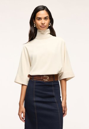 Donna con dolcevita bianco a maniche corte larghe, cintura marrone, gonna blu scuro a vita alta e orecchini a cerchio dorati su sfondo uniforme.