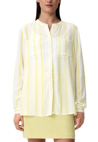 Camicia bianca con righe verticali gialle e azzurre chiare, maniche lunghe e due taschini sul petto, abbinata a una gonna giallo chiaro.
