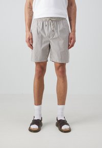 Uomo che indossa pantaloni corti verticali a righe beige e neri, calze bianche alla caviglia e sandali in pelle marrone scuro, in piedi davanti a uno sfondo bianco semplice.