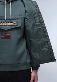Felpa con cappuccio verde scuro, dotata di una fodera interna con zip, caratterizzata da un patch del logo e da un piccolo patch della bandiera norvegese sulla parte frontale.
