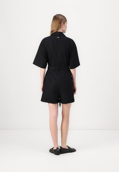 Combinaison noire à manches courtes avec une taille ceinturée, dotée d'un col et d'une étiquette près du cou. Tissu lisse ; coupe décontractée. Chaussures noires.