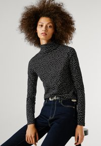 Göndör hajú nő fekete, hosszú ujjú garbóban, fehér pöttyökkel, sötét farmerben ül egy széken, sima háttér előtt.