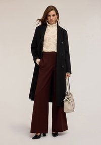Palton negru din lână peste un pulover tricotat crem, asortat cu pantaloni burgundii cu talie înaltă și pantofi cu toc din piele lăcuită negri. Accesorizat cu o geantă bej.