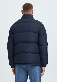 Marineblå puffjakke med høj krave, quiltet design og elastiske manchetter. Båret sammen med lyseblå jeans. Standard pasform, afslappet stil.