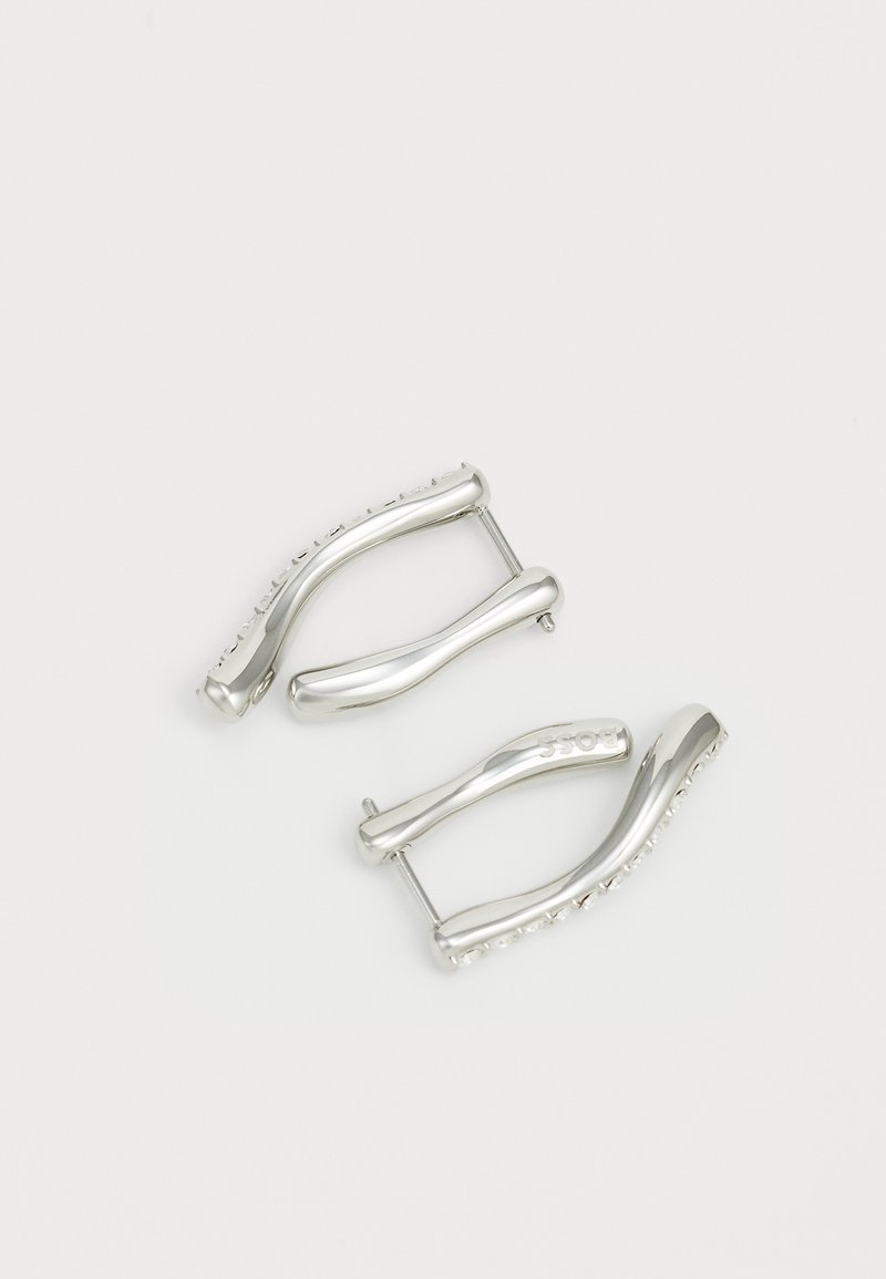 Pendientes de metal plateado con un diseño en forma de ola, que presentan curvas suaves y un borde texturizado. Primer plano del detalle del herraje visible.