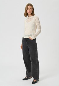 Bluză cu mâneci lungi din dantelă, de culoare crem, cu un design ajustat, purtată cu blugi negri cu talie înaltă și picioare largi, și balerini negri cu vârful ascuțit.