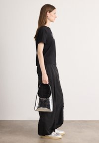 T-shirt noir court, pantalons noirs amples, tenant un sac à main métallique argenté, associé à des baskets de couleur claire. Fond uni.