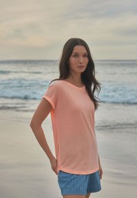 Heledad roosa lühikeste varrukatega T-särk �ümarate kaelustega, koos siniste ja valgete triibuliste lühikeste pükstega. Pehme tekstuur; mugav lõike. Ranna taust.