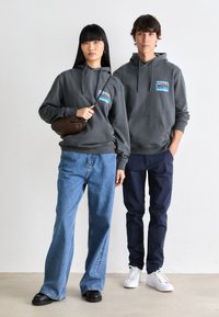 Grijze hoodies met voorzakken, voorzien van een kleurrafisch. Model links in blauwe wijdere jeans; model rechts in marineblauwe broek en witte sneakers.