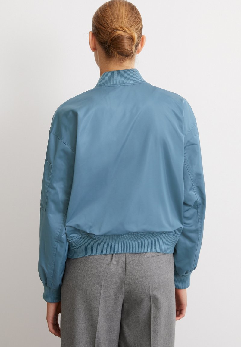 Jasnoniebieska kurtka bomberka z prążkowanym kołnierzem i mankietami. Gładki, błyszczący materiał o luźnym kroju, pokazana od tyłu, zestawiona z szarymi spodniami.