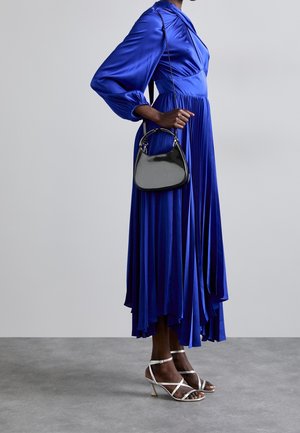 Personne portant une longue robe plissée bleu roi, tenant un petit sac à main noir et portant des sandales à talons blanches à lanières, debout sur un sol gris.