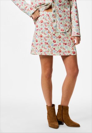 Žena stojaca na bielom pozadí, oblečená v súprave kvetinovo vzorovanej sukne a košele, s hnedými nubukovými členkovými čižmami.