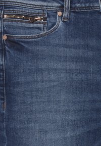 Jeans de denim en azul oscuro, con un bolsillo frontal con cremallera y herrajes metálicos, y una textura suave con un ligero desgastado.