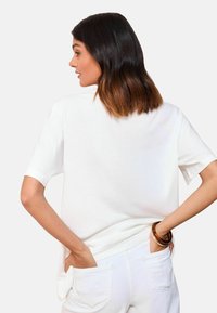 Weißes Kurzarm-T-Shirt mit lockerer Passform, aus weichem Baumwollmaterial. Der Rücken hat einen runden Ausschnitt und es wird mit leichten weißen Hosen kombiniert.
