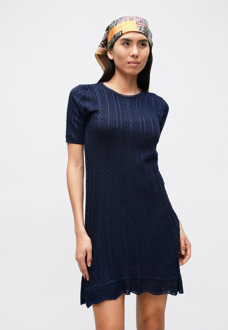 Navy blue knitted dress with short sleeves, textured zigzag pattern, scalloped hem, and a round neckline. Accented with a patterned headscarf.