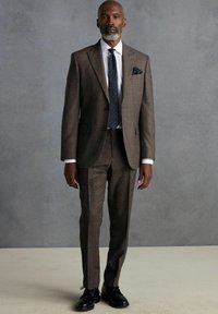 Brauner Tweed-Anzug mit strukturiertem Finish, weißem Hemd und navyblauer Krawatte mit Punkten. Auffälliges Taschentuch-Detail, ergänzt durch schwarze Schuhe.
