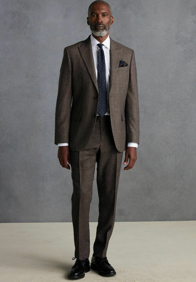 Costume en tweed marron avec une finition texturée, chemise blanche et cravate à pois bleus marins. Détail de pochette notable, complété par des chaussures noires.