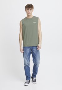 Ærmeløs olivengrøn t-shirt med "A.R.C." logo, parret med lysblå denim jeans og sorte højtop sneakers. Ensfarvet baggrund.