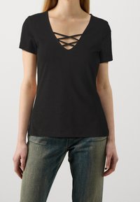 Schwarzes T-Shirt mit kurzen Ärmeln und V-Ausschnitt, der mit Kreuzdetails verziert ist. Hergestellt aus weichem Stoff mit einer taillierten Silhouette, kombiniert mit hellblauen Jeans.