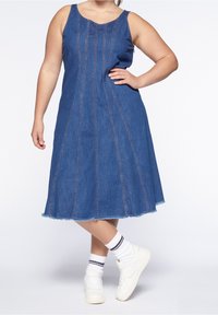 Jeansklänning med en figurnära överdel, utsvängd kjol och rå fåll. Djupblå färg med kontrasterande sömmar. Bärs med vita sneakers.