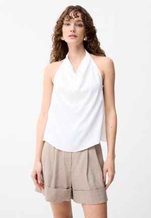 Vrouw met krullend haar, draagt gouden oorbellen, een witte haltertop en beige opgerolde korte broek, staand tegen een eentonige achtergrond.